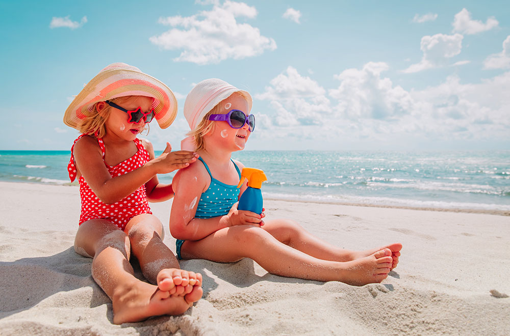 Duas meninas na praia, de fato de banho, óculos de proteção e chapéu, a aplicar protetor solar.