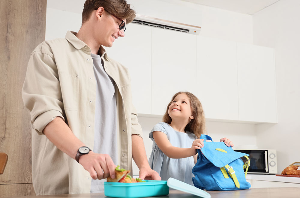 O pai está a preparar o almoço para a filha, que sorri enquanto segura a mochila.