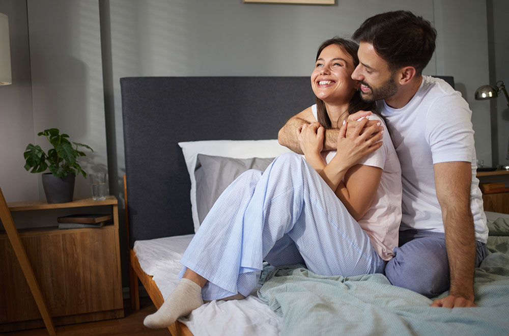 Um casal rindo e se abraçando na cama, compartilhando um momento aconchegante e feliz juntos.