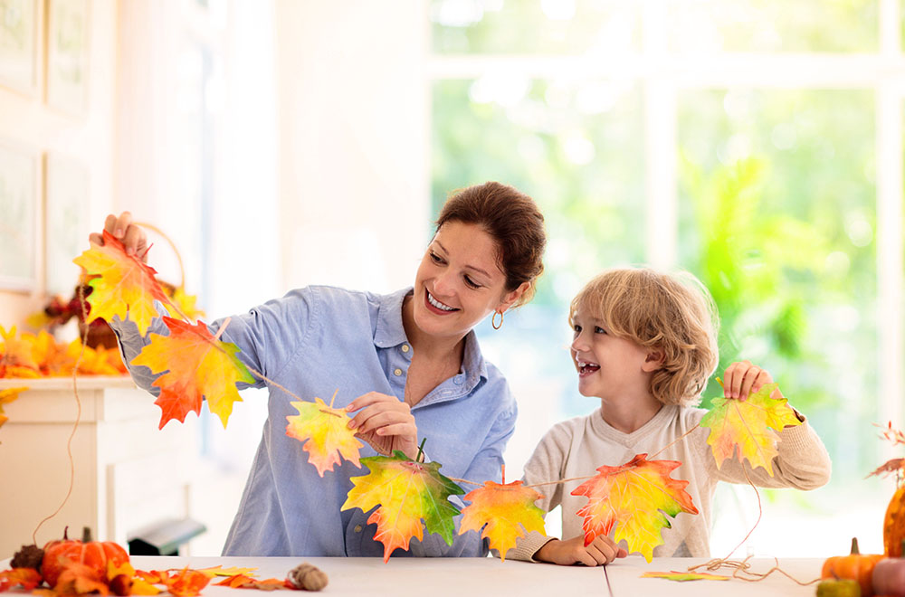 Mãe e filho fazem uma guirlanda de folhas coloridas, sorrindo um para o outro em uma sala clara e ensolarada.