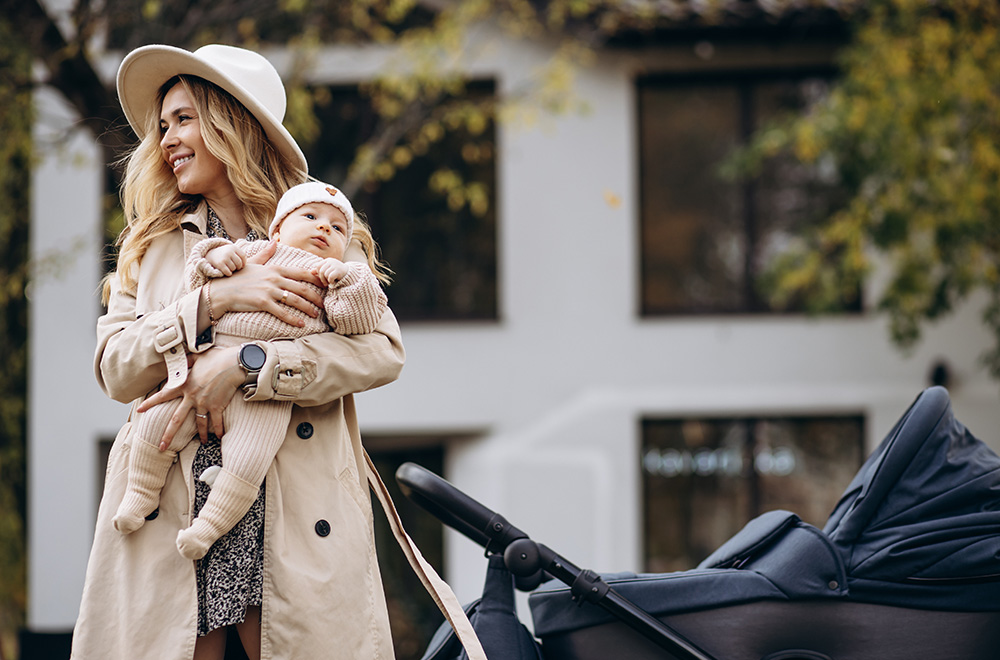 Uma mulher segura um bebê nos braços enquanto caminha ao lado de um carrinho em frente a um edifício.